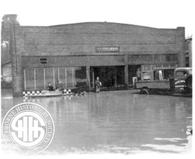 Historic Hawthorne Post Office building affected by flooding, with water reaching the entrance and vintage vehicles parked outside, showcasing early 20th-century architecture.