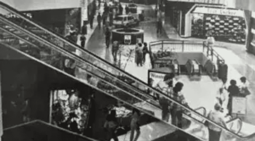 Historic Hawthorne shopping mall escalator scene, showing shoppers and storefronts in black and white.