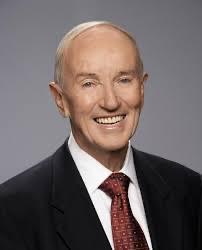 A portrait of an elderly man with a friendly smile, dressed in a dark suit and red tie, representing community history and local heritage.