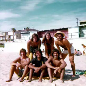 Beach group photo of six people enjoying a sunny day on the sand, smiling and posing together, with colorful buildings and other beachgoers in the background.