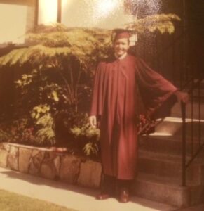 Woman in maroon graduation gown standing outdoors on steps with greenery in background.