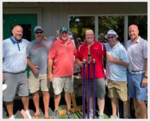 Group of men at a community event, standing outdoors in front of a building, smiling and enjoying the summer day.