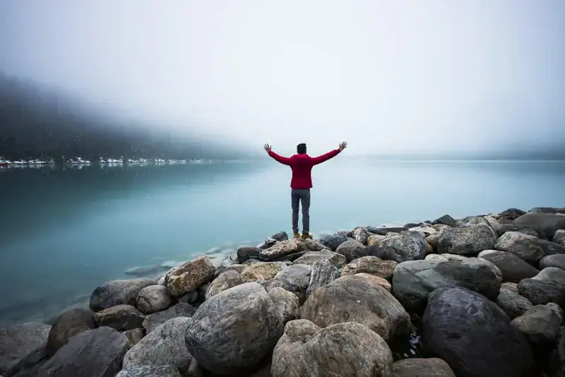 A person standing on rocks by a calm lake with arms outstretched, symbolizing faith and love for God amidst serene natural surroundings.