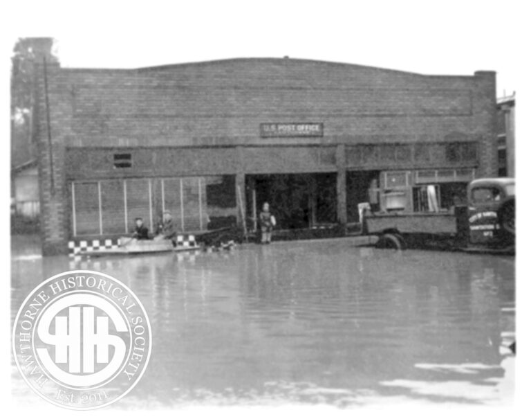 Historic Hawthorne Post Office building affected by flooding, with water reaching the entrance and vintage vehicles parked outside, showcasing early 20th-century architecture.