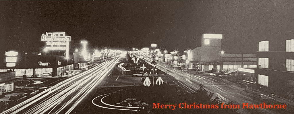 Night view of Hawthorne's Main Street with light trails from moving vehicles, illuminated storefronts, and festive holiday lights.
