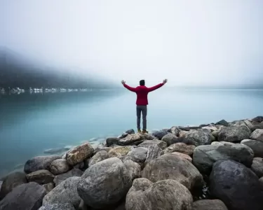 A person standing on rocks by a calm lake with arms outstretched, symbolizing faith and love for God amidst serene natural surroundings.