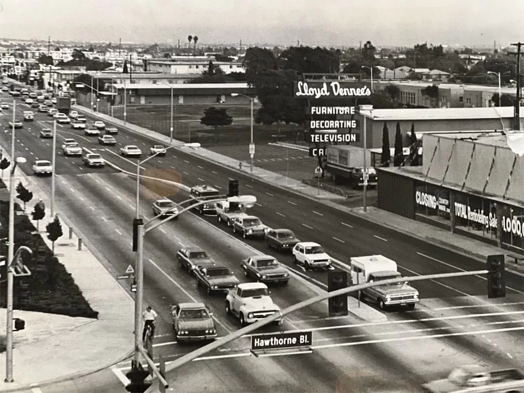 Historic black and white photo of Hawthorne Boulevard showing vintage cars and storefronts, including Lloyd Dennes furniture and decor store, capturing mid-20th-century commercial life.